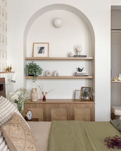 Arched niche with wooden shelves, decor objects, and cane-front cabinet above a bed with green quilt