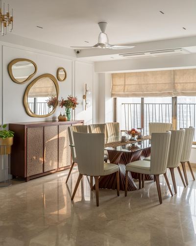 Dining room with glass table on carved wood pedestal, cream chairs, and cluster of round mirrors above sideboard