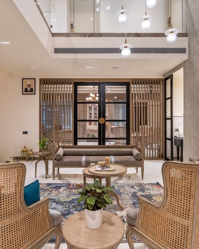 Double-height living room with cane armchairs, marble floor, and fluted wood screens framing black doors