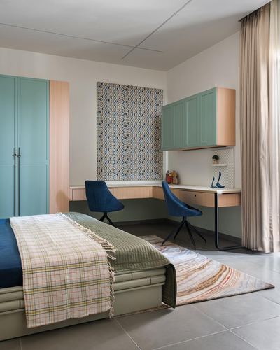 Bedroom corner with teal wardrobes, built-in desk, two blue chairs, and layered bedding on rug