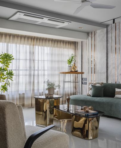 Living room with green velvet sofa, gold sculptural tables, and marble-clad wall behind sheer curtained window