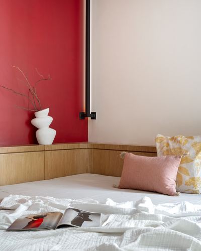 Bed corner with red accent wall, wooden headboard nook, stacked white vase and floral cushions
