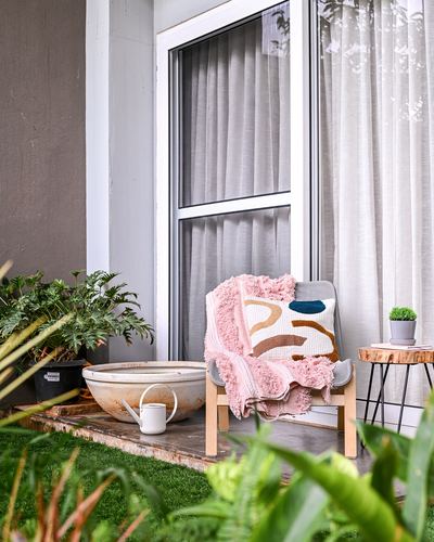 Small balcony seating nook with wooden armchair, pink throw, patterned cushion, and potted plants beside a curtained glass door