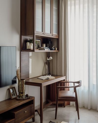 Wooden study nook with glass-front cabinet, open shelf, desk, lamp, and leather armchair by sheer-curtained window