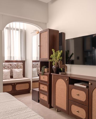 Bedroom corner with arched window seat, cane wardrobe, mirror, and wood console with wall-mounted TV