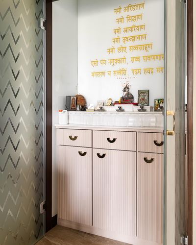 Pooja cabinet with fluted pink doors, brass handles, idols, and gold Jain prayer text on glass