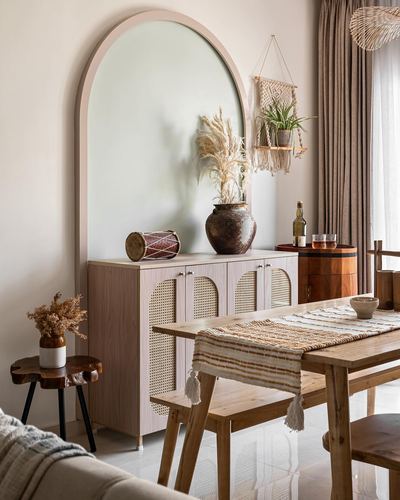 Dining nook with arched mirror above cane-front cabinet and wooden table in Japandi-style apartment