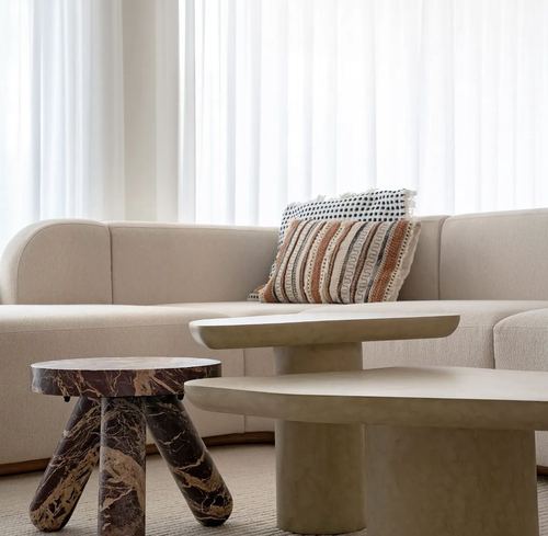 Beige curved sofa with striped cushion beside layered stone coffee tables near sheer curtains