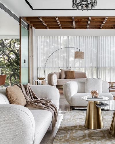 Curved cream sofas around brass coffee tables beneath a wood-coffered ceiling with sheer curtained glass walls