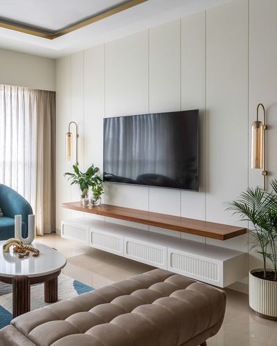 White paneled living room with wall-mounted TV, floating wood shelf, brass sconces, and tufted beige ottoman