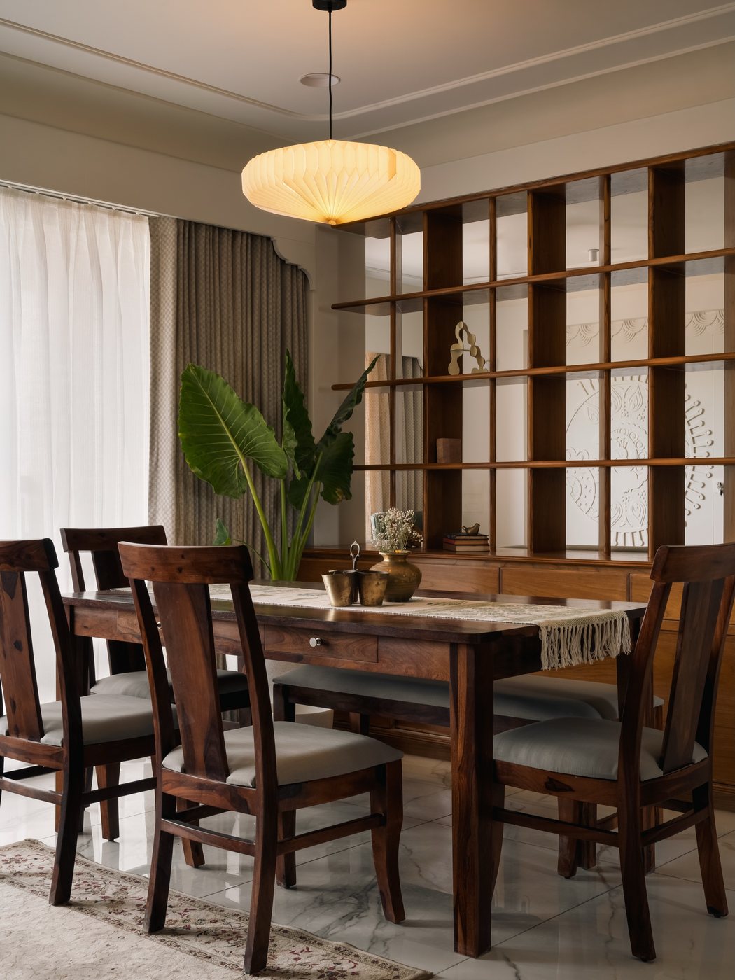 Wooden dining table with six chairs beneath a glowing pendant light, jali shelving behind
