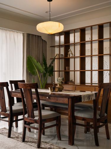 Wooden dining table with six chairs beneath a glowing pendant light, jali shelving behind