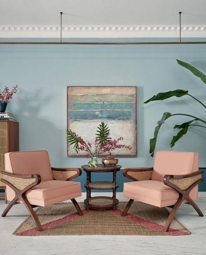 Two peach cane-armed chairs flank a round wooden table under an abstract painting on a blue wall