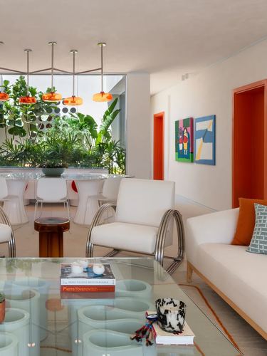 Living area with chrome armchair, glass table, and orange pendant lights over a dining table framed by greenery