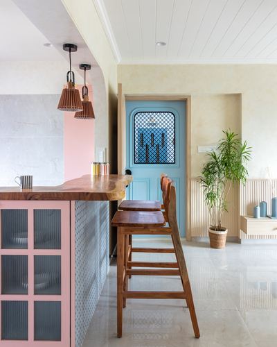 Pastel pink kitchen island with wooden stools facing a blue arched door and plant.