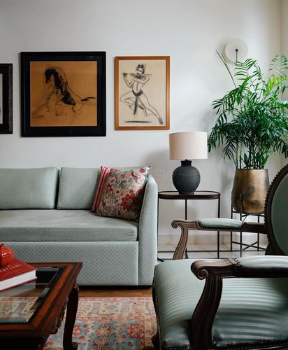 Sage sofa with floral cushion beneath framed sketches, brass planter and striped armchair nearby