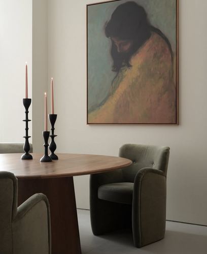 Round wood dining table with black candlesticks beneath a large framed portrait painting, olive velvet chairs