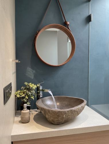 Round leather-strap mirror above a carved stone vessel sink against a blue textured wall