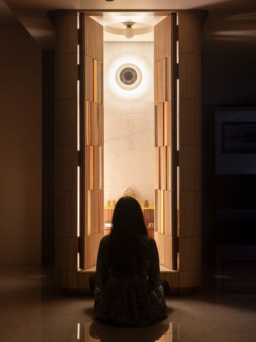 Woman seated before a cylindrical travertine temple niche with backlit wooden doors and a glowing marble interior