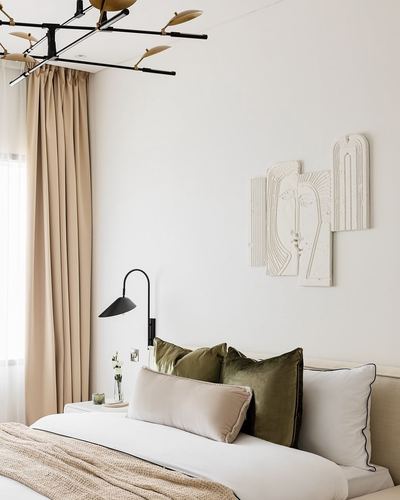 Neutral bedroom with upholstered headboard, olive cushions, sculptural wall art, and brass-and-black chandelier