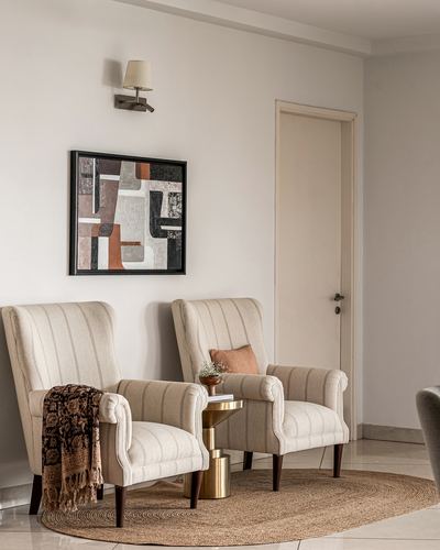 Two beige wingback armchairs beside a brass side table under abstract art on a jute rug