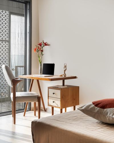 Sunlit corner with wooden desk, cane chair, and bedside drawers beside a bed