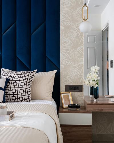Bedroom corner with navy tufted headboard, patterned pillows, and wood nightstand with vase and pendant light