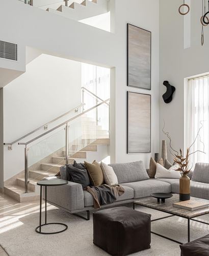 Neutral-toned living room with sectional sofa beneath tall artwork, glass-railed staircase beside double-height wall