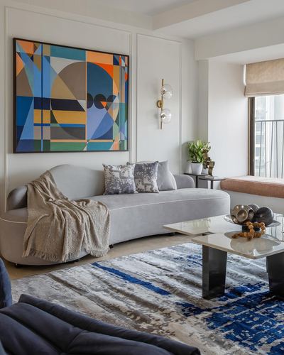 Grey curved sofa with patterned cushions beneath a colorful geometric painting, blue-patterned rug and marble coffee table.
