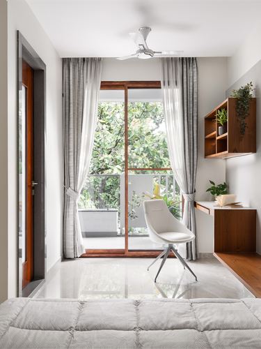 Small study nook with wood desk, white chair, and glass door opening to a leafy balcony
