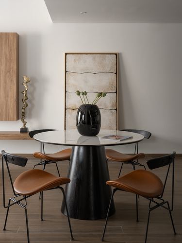Round marble dining table with black conical base and tan leather chairs against a leaning stone artwork