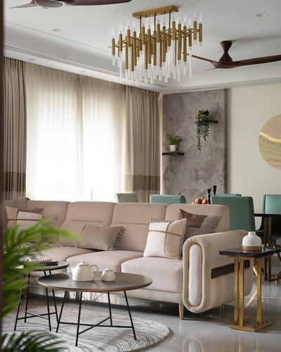 Beige sectional sofa under brass chandelier with marble accent wall and green dining chairs beyond