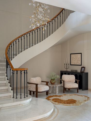Curved marble staircase with wrought iron railing beside a seating nook with two patterned armchairs.