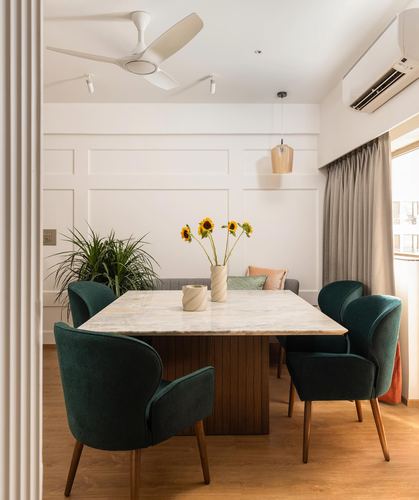 Dining room with marble table, green velvet chairs, sunflowers, and paneled white walls