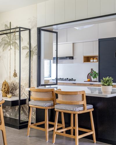 Kitchen island with cane barstools seen through black-framed folding glass door, pooja nook at left