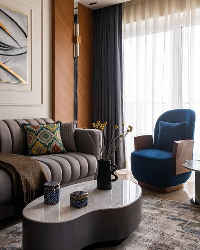 Living room corner with grey channel-tufted sofa, navy velvet armchair, and curved marble-top coffee table beside sheer curtains.