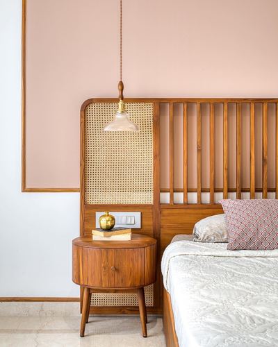 Teak and cane headboard beside a round wooden nightstand under a pendant light, pink wall behind.