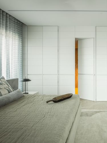 Minimalist bedroom with grey bedding, paneled white wardrobe wall, and a glowing ajar door