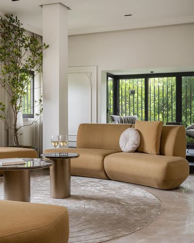 Curved mustard sofa on round rug beside marble floor, glass wall to greenery