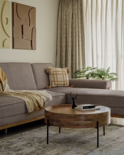 Taupe L-shaped sofa with mustard throw beside round wooden coffee table and abstract wall art