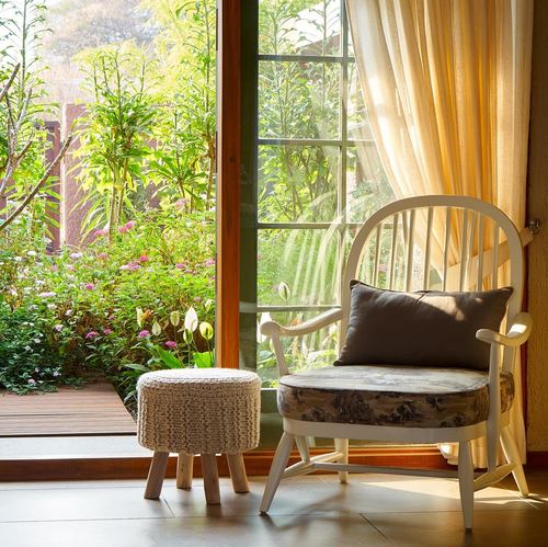 White windsor armchair and knit pouf beside a large window overlooking a lush garden