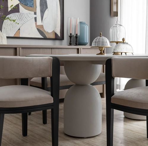 Dining table with sculptural pedestal base, beige velvet chairs, and abstract artwork behind sideboard