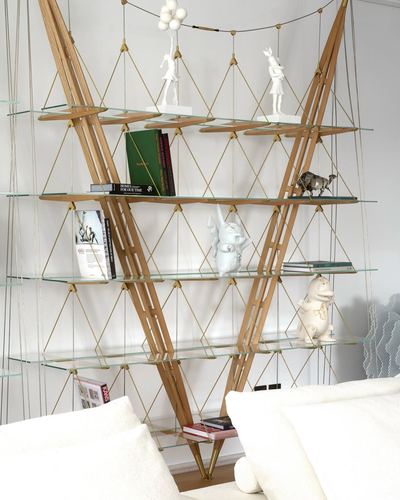 Sculptural wooden bookshelf with brass cables and glass shelves holding books and figurines, boucle sofa in front
