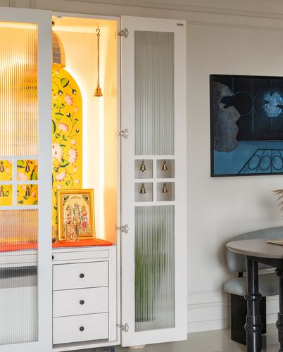 White built-in pooja cabinet with reeded glass doors and glowing yellow floral niche holding deity idols