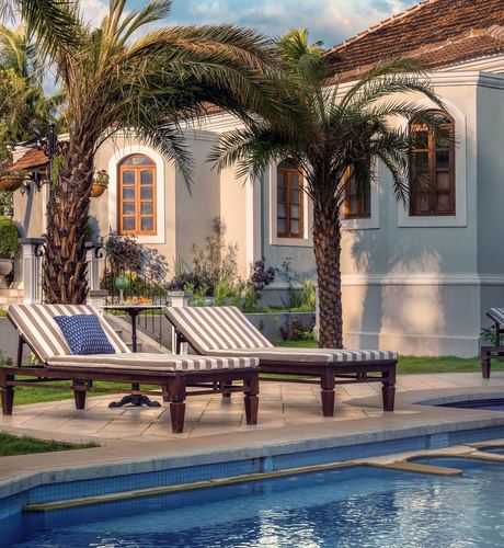 Poolside loungers under palm trees facing a cream colonial mansion with arched windows in Goa