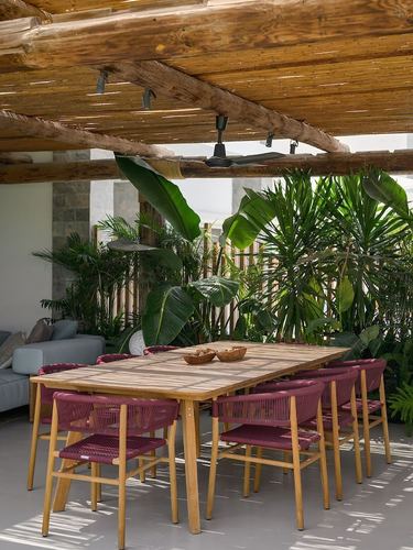 Outdoor dining table under a wooden pergola with burgundy rope chairs and lush plants behind.