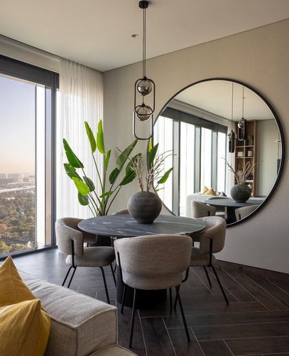 Dining nook with round marble table, cane chairs, and large circular mirror against floor-to-ceiling window with city view