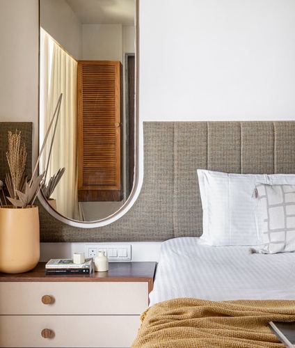 Bedroom corner with channel-tufted headboard, oval mirror reflecting louvered wardrobe, and wooden bedside chest