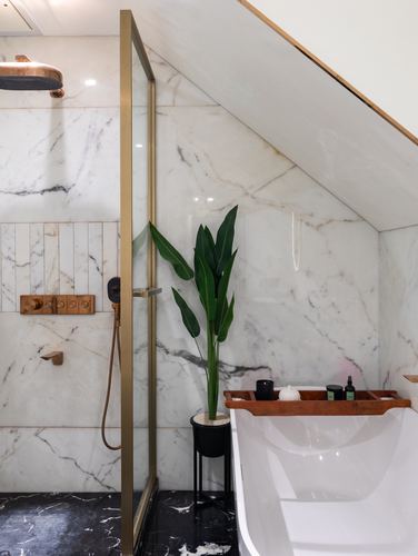 Marble-clad bathroom with brass shower fittings, glass partition, freestanding tub, and a potted plant under a sloped ceiling