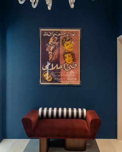 Rust velvet curved bench with striped bolster under a framed vintage Arabic film poster on a deep blue wall.